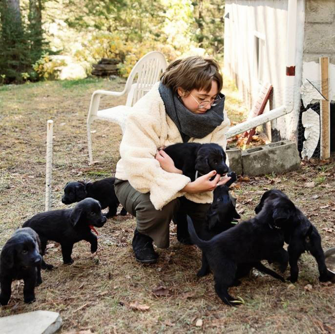 A woman, kneeling down outside, is surrounded by black puppies