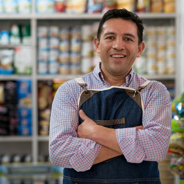 Un propriétaire de petite entreprise se tient debout dans son épicerie, les bras croisés sur la poitrine et un grand sourire sur le visage. Derrière lui se trouvent des étagères remplies de provisions.