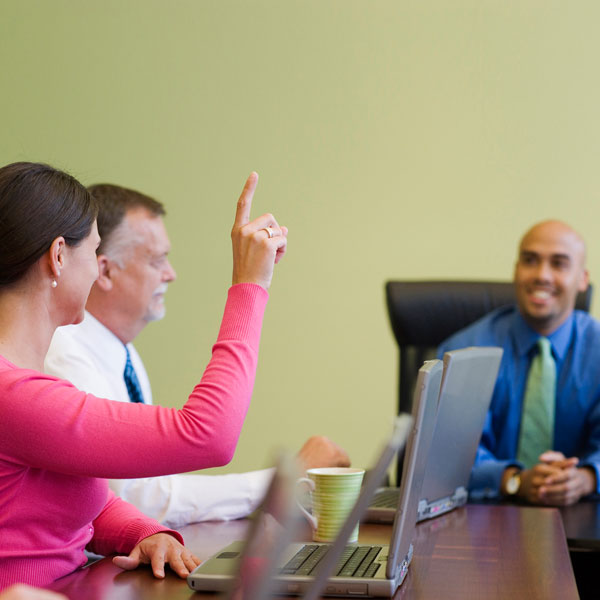 3 people are sitting at a conference table. One woman has her hand up to ask a question.